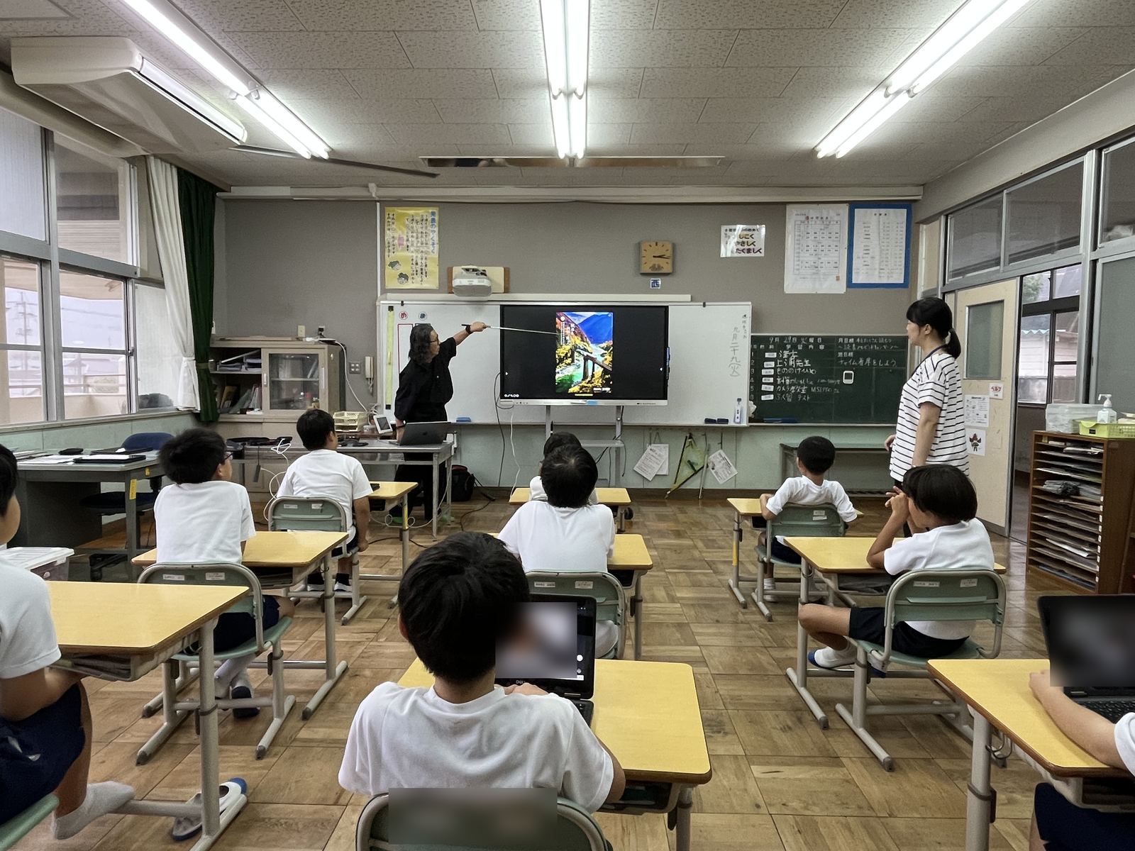 モニターに映した風景写真を指し棒で示しながら説明する講師と、それを見つめる児童たち