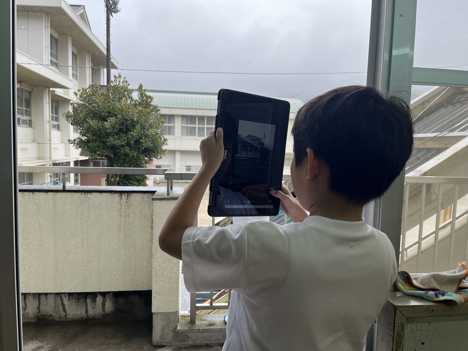タブレットを縦に構えて、窓の外の木と校舎を撮影する児童の後ろ姿
